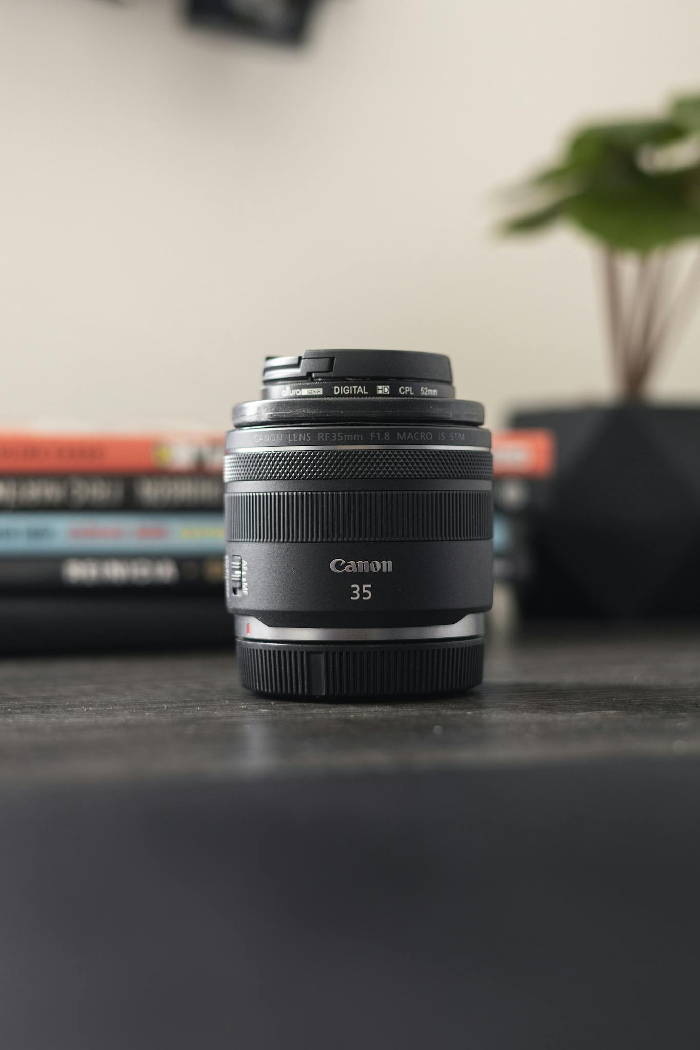 A Canon 35mm lens sits in focus on a desk with books and a plant in the background.